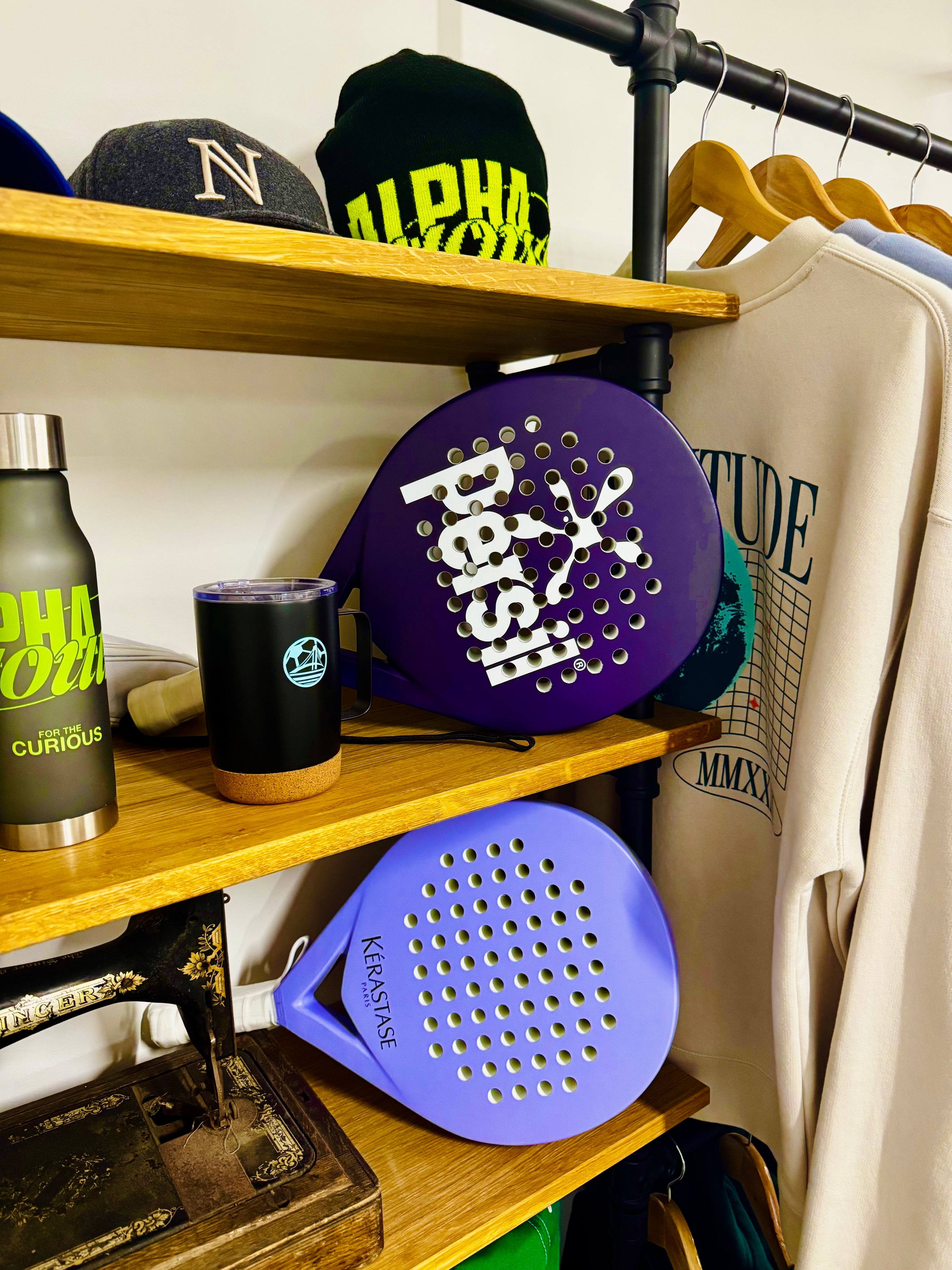 A styled retail display featuring two purple paddle rackets on wooden shelves, alongside a black tumbler, a Curious Curated bottle, and a cream sweatshirt hanging on a rack.
